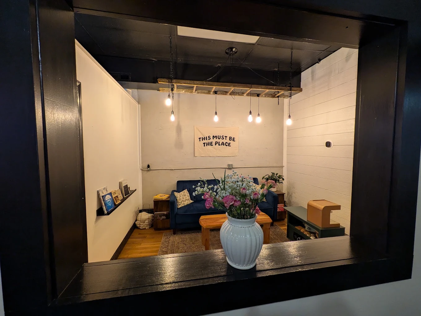 The back lounge seen through an interior window with a vase of flowers on the sill, and a banner reading This Must Be The Place.