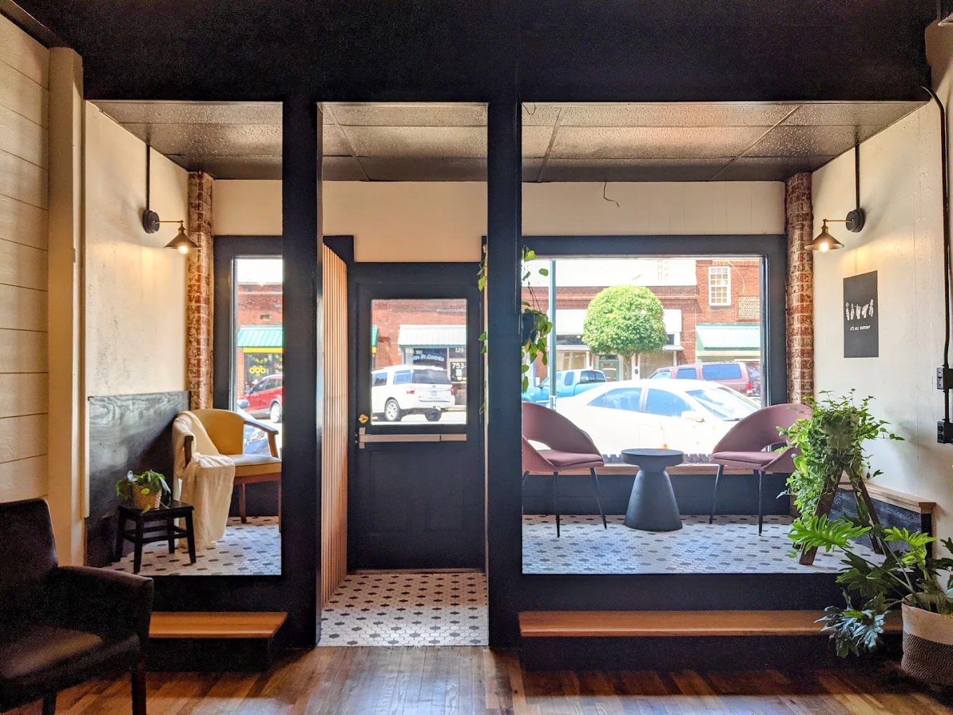 Two window nooks at the front of the shop, with pink chairs on white hex tile, hanging plants and Main Street beyond the glass.