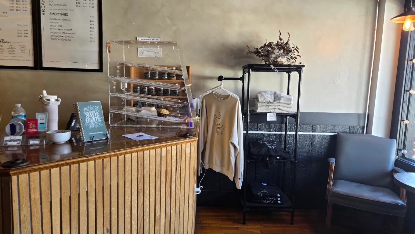 A cream crewneck sweatshirt with a line drawing of stacked coffee cups hangs on a pipe rack, beside shelves of folded white and black sweatshirts.